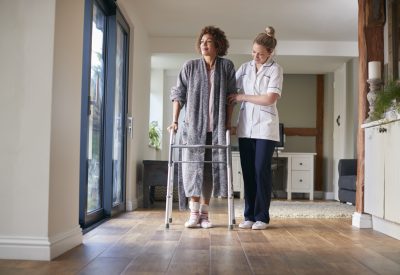 Mature Woman In Dressing Gown Using Walking Frame Being Helped By Female Nurse
